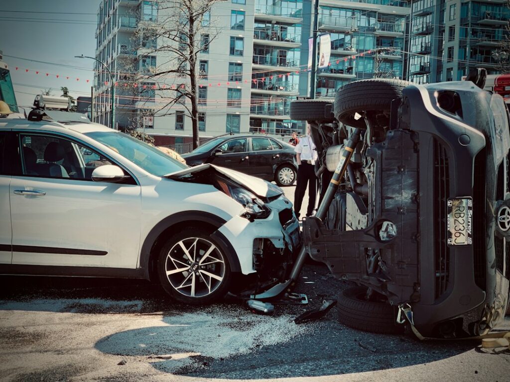 Two drivers stand beside damaged vehicles while exchanging information and photographing the crash scene.